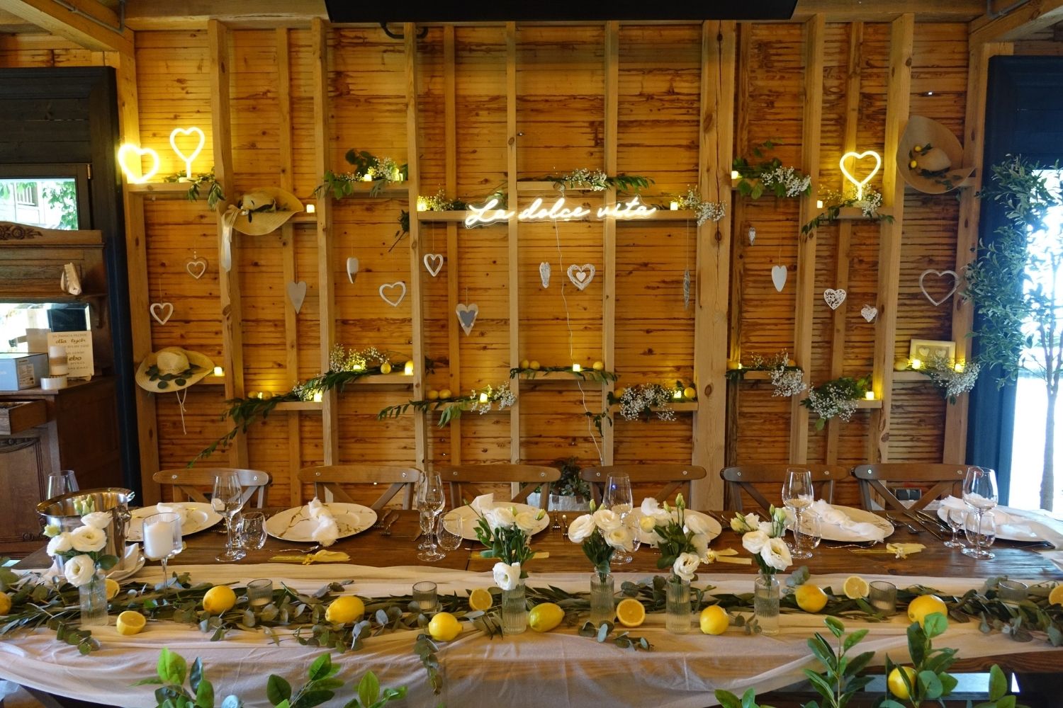 Dekoracja stołu weselnego w stylu włoskim, drewniany stół z girlandą z cytryn i neonem La dolce vita na drewnianej ścianie.