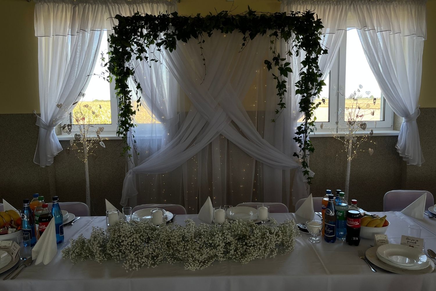 Ścianka weselna za stołem pary młodej, białe zasłony z zieloną girlandą i gipsówka na stole.