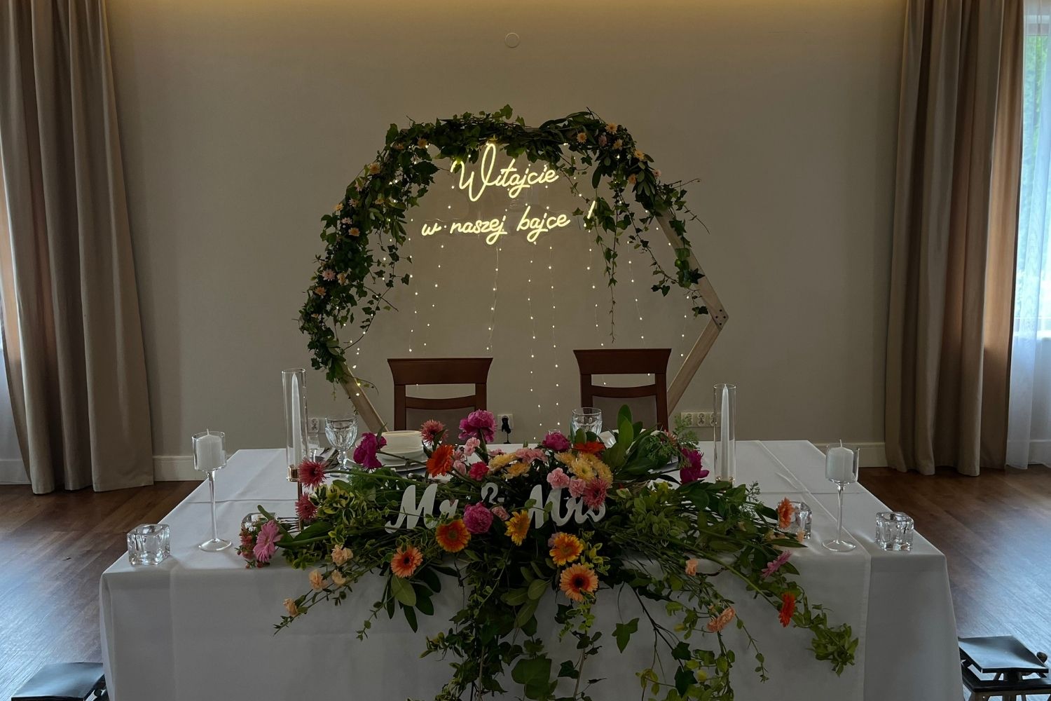 Heksagonalna ścianka weselna z neonem "Witajcie w naszej bajce" i kompozycją kwiatową na stole pary młodej.