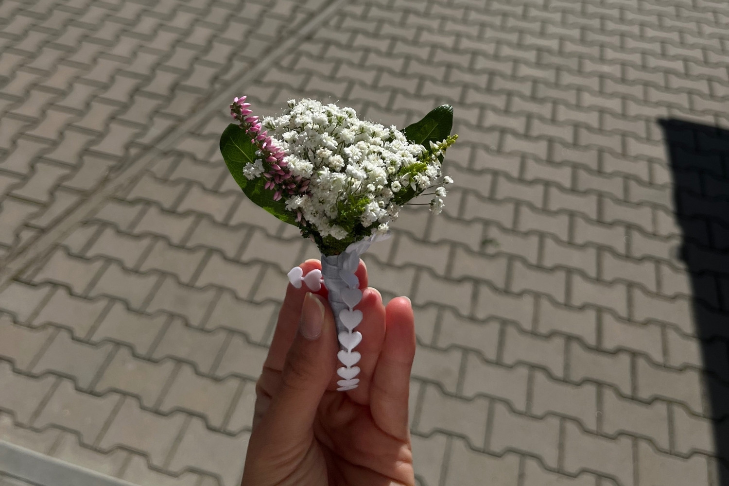 Mała butonierka ślubna z gipsówki i wrzosu, z ozdobną białą wstążką w kształcie serc.